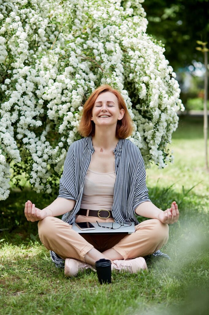 Happy woman meditates while working remotely in the park with laptop and phone among the greenery